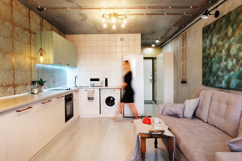 На фото: угловая, светлая кухня-гостиная в стиле лофт с накладной мойкой, плоскими фасадами, белыми фасадами, разноцветным фартуком, черной техникой, бежевым полом, серой столешницей и диваном без острова