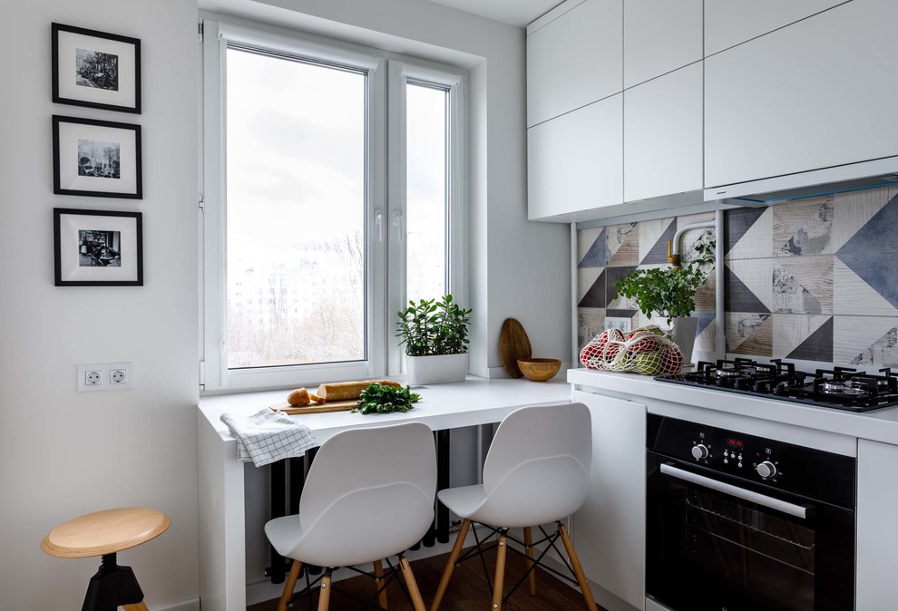 Aménagement d'une cuisine scandinave fermée avec un placard à porte plane, des portes de placard blanches, une crédence multicolore, un électroménager noir et un plan de travail blanc.