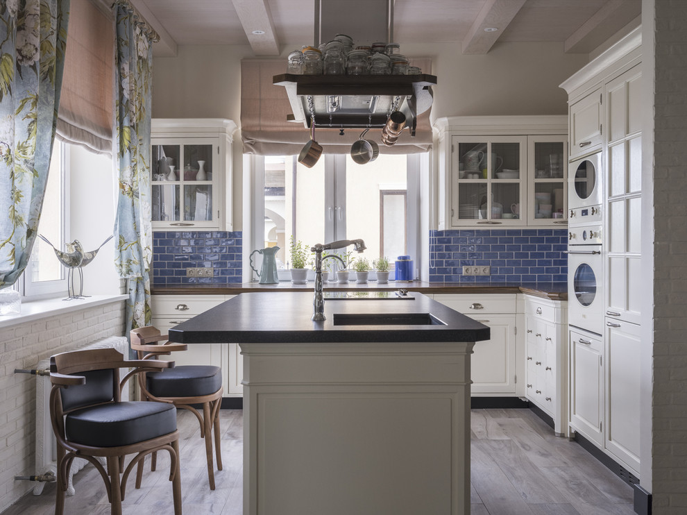 Пример оригинального дизайна: п-образная кухня-гостиная в классическом стиле с врезной мойкой, фасадами с утопленной филенкой, белыми фасадами, синим фартуком, белой техникой, островом, серым полом и коричневой столешницей