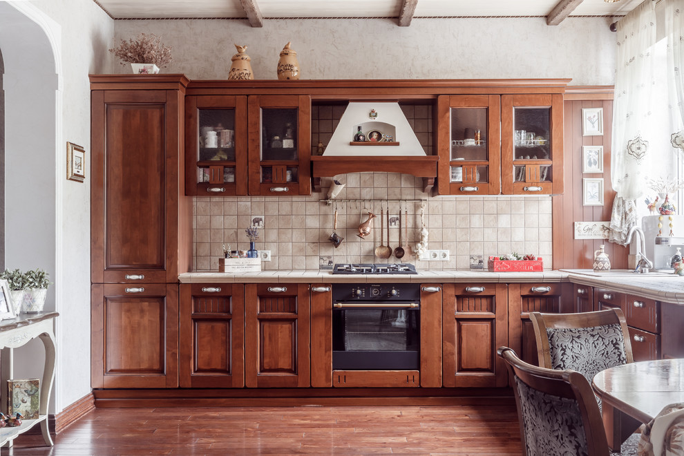 Exempel på ett klassiskt beige beige kök, med kaklad bänkskiva, en nedsänkt diskho, luckor med upphöjd panel, skåp i mellenmörkt trä, beige stänkskydd, svarta vitvaror och brunt golv