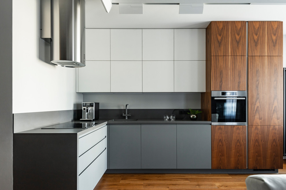 Пример оригинального дизайна: угловая кухня-гостиная в современном стиле с одинарной мойкой, плоскими фасадами, серыми фасадами, паркетным полом среднего тона, черной столешницей и двухцветным гарнитуром