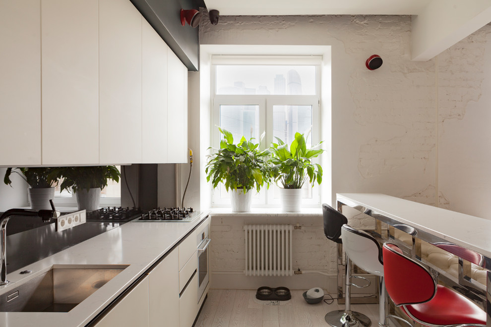 Свежая идея для дизайна: прямая кухня-гостиная в современном стиле с врезной мойкой, плоскими фасадами, белыми фасадами, черным фартуком, белой техникой, светлым паркетным полом и бежевым полом - отличное фото интерьера