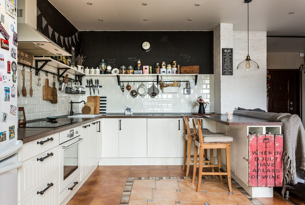Inredning av ett modernt kök, med en nedsänkt diskho, vita skåp, träbänkskiva, vitt stänkskydd, en halv köksö, brunt golv, stänkskydd i tunnelbanekakel och vita vitvaror