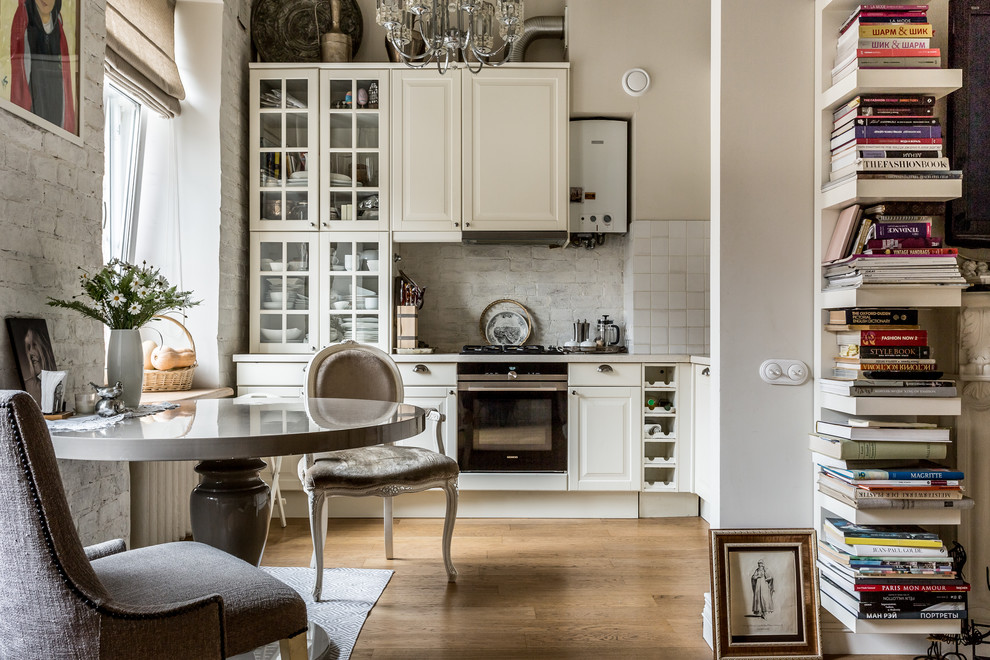 На фото: угловая кухня-гостиная в современном стиле с фасадами с выступающей филенкой, белыми фасадами, белым фартуком, черной техникой и коричневым полом