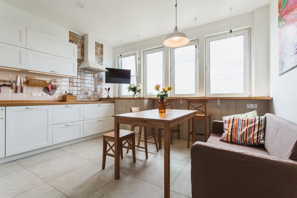 На фото: кухня в скандинавском стиле с фасадами в стиле шейкер, белыми фасадами, деревянной столешницей, разноцветным фартуком и диваном без острова с