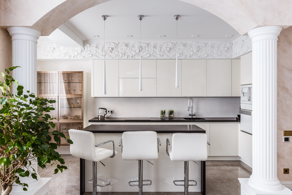 Пример оригинального дизайна: угловая кухня-гостиная в стиле неоклассика (современная классика) с плоскими фасадами, белыми фасадами, белым фартуком, белой техникой, островом, бежевым полом и черной столешницей