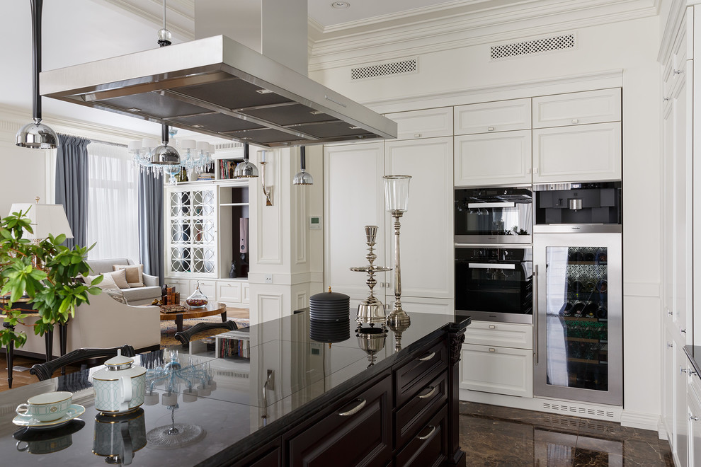 На фото: кухня-гостиная среднего размера в стиле неоклассика (современная классика) с мраморным полом, коричневым полом, белыми фасадами, островом, фасадами с утопленной филенкой и черной техникой