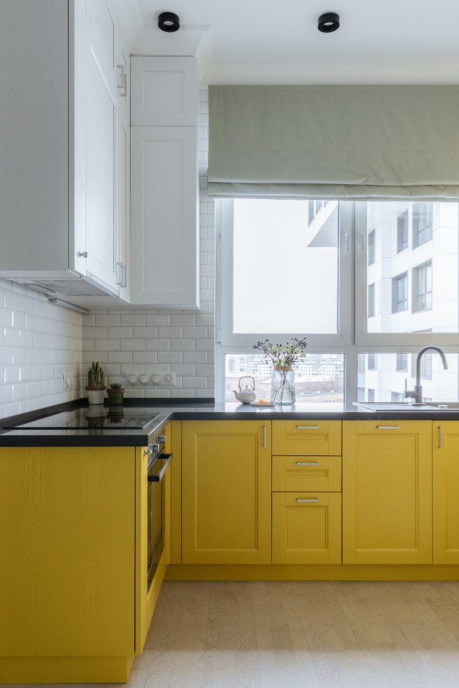 На фото: угловая кухня в стиле неоклассика (современная классика) с накладной мойкой, фасадами с утопленной филенкой, желтыми фасадами, белым фартуком, фартуком из плитки кабанчик, черной техникой, светлым паркетным полом, бежевым полом, черной столешницей и двухцветным гарнитуром с
