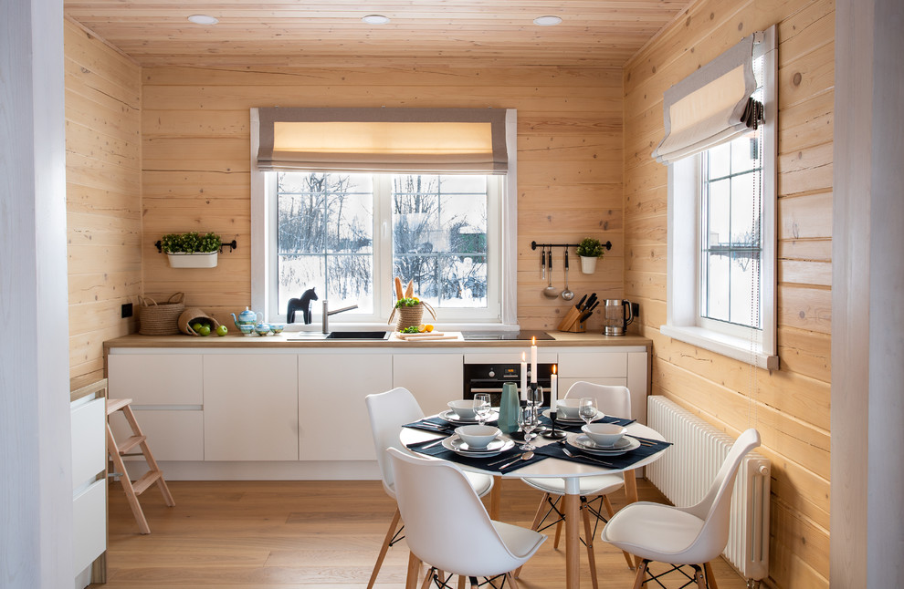 На фото: прямая, светлая кухня в скандинавском стиле с обеденным столом, накладной мойкой, плоскими фасадами, белыми фасадами, деревянной столешницей, черной техникой, паркетным полом среднего тона, коричневым полом и коричневой столешницей