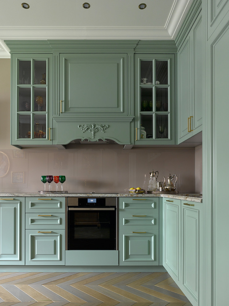 Inredning av ett klassiskt beige beige l-kök, med luckor med upphöjd panel, gröna skåp, svarta vitvaror, mellanmörkt trägolv, brunt stänkskydd, glaspanel som stänkskydd och brunt golv