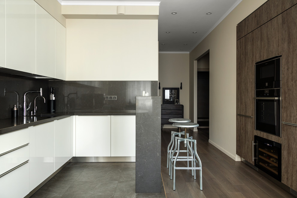На фото: угловая кухня в современном стиле с плоскими фасадами, белыми фасадами, серым фартуком, черной техникой, серым полом и черной столешницей