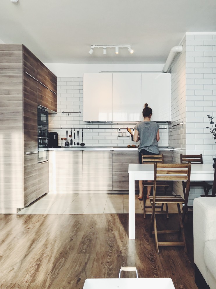 This is an example of a medium sized scandinavian l-shaped open plan kitchen in Moscow with a submerged sink, flat-panel cabinets, medium wood cabinets, white splashback, metro tiled splashback, stainless steel appliances, laminate floors, no island, beige floors and white worktops.