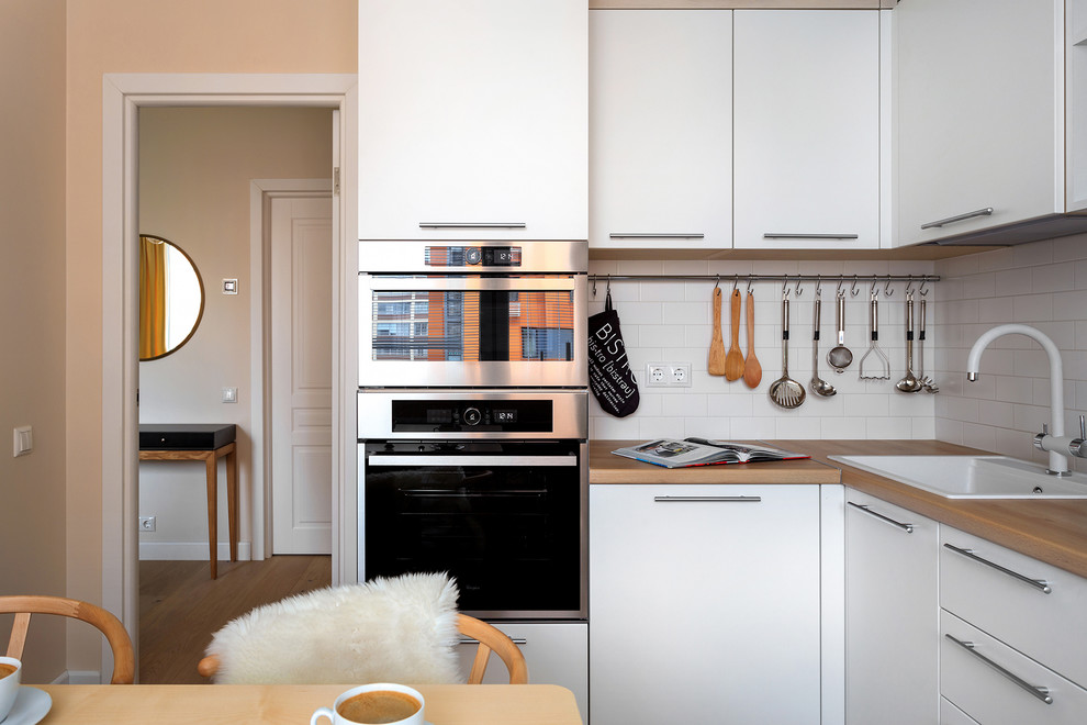 Small scandinavian l-shaped enclosed kitchen in Moscow with a built-in sink, flat-panel cabinets, white cabinets, laminate countertops, white splashback, ceramic splashback, stainless steel appliances, light hardwood flooring and beige floors.