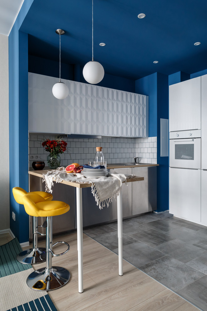 Bild på ett funkis l-kök, med en nedsänkt diskho, släta luckor, skåp i rostfritt stål, träbänkskiva, vitt stänkskydd, vita vitvaror, en halv köksö och grått golv