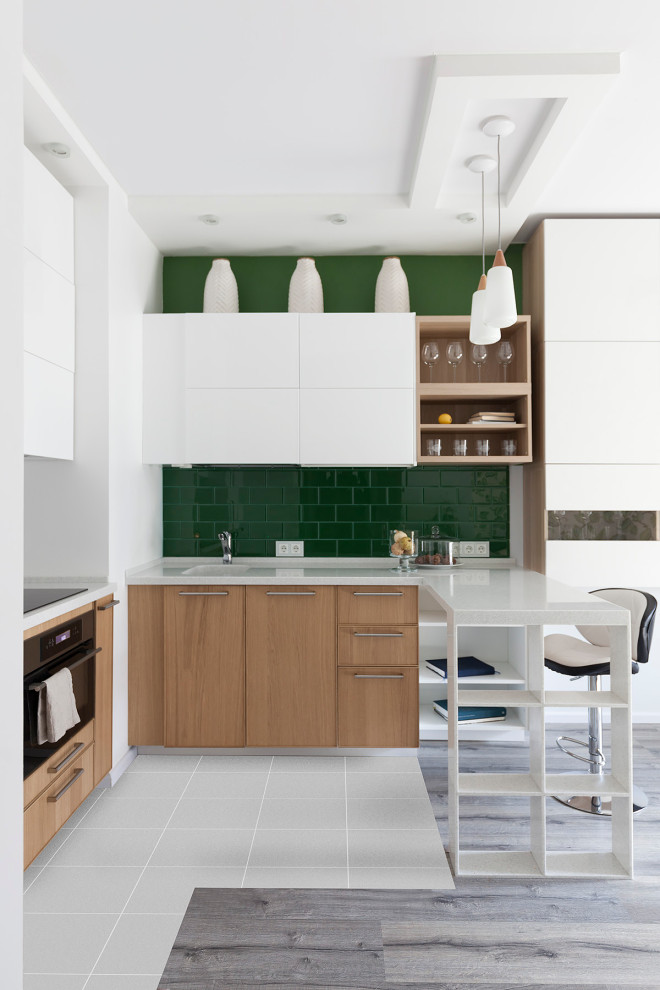 На фото: п-образная, светлая кухня в современном стиле с одинарной мойкой, плоскими фасадами, белыми фасадами, зеленым фартуком, фартуком из плитки кабанчик, полуостровом, серым полом и белой столешницей