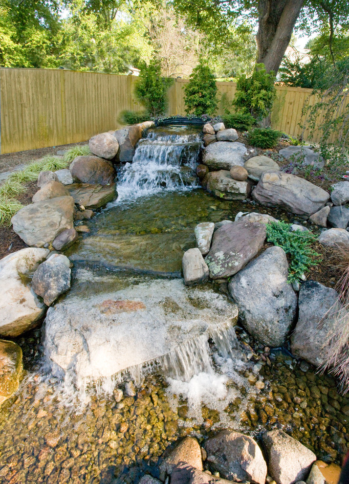 Imagen de jardín clásico grande en verano en patio trasero con fuente, exposición total al sol y adoquines de piedra natural