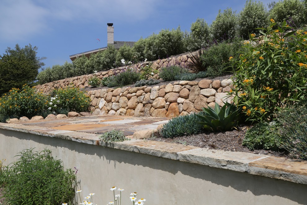Diseño de jardín mediterráneo en patio lateral con muro de contención, exposición total al sol y adoquines de piedra natural