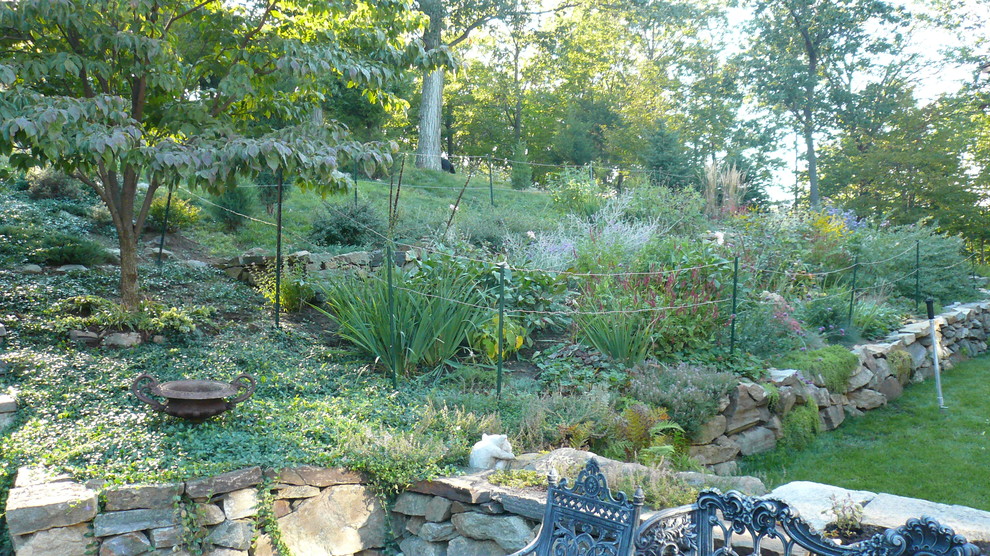Inredning av en rustik stor trädgård i full sol längs med huset på sommaren, med en stödmur och naturstensplattor