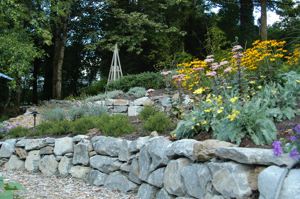 На фото: большой регулярный сад на склоне в классическом стиле с мульчированием, подпорной стенкой и полуденной тенью с