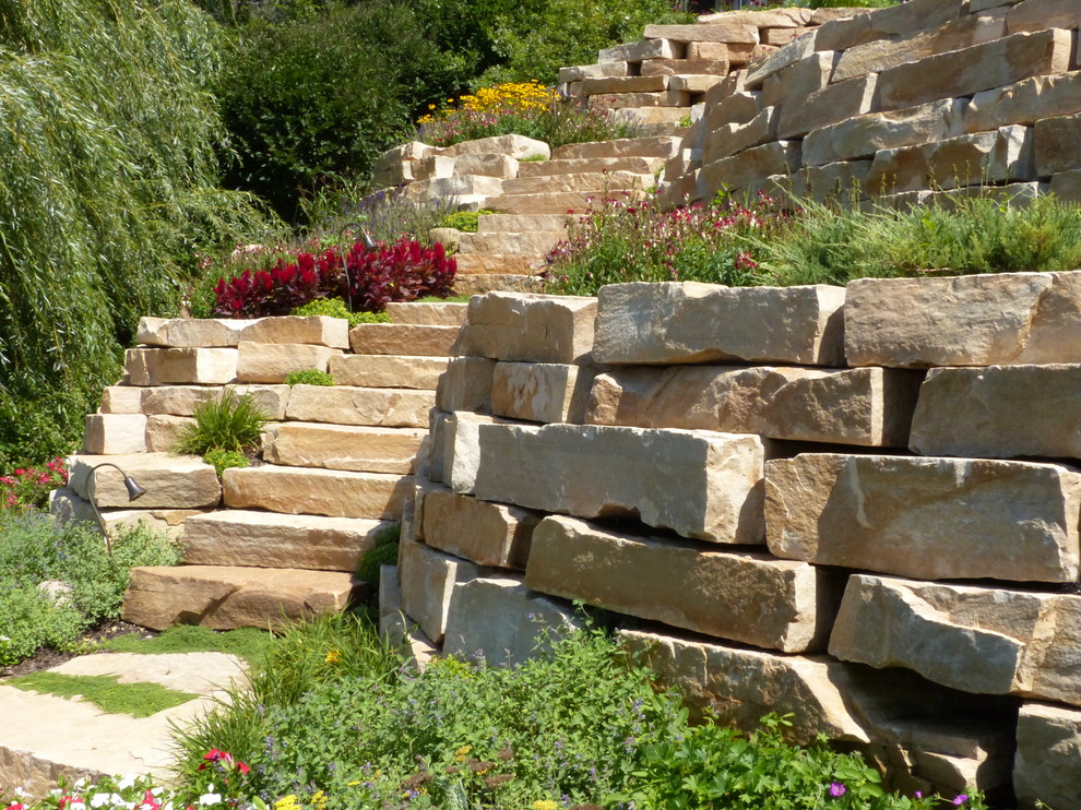 Modelo de jardín tradicional renovado de tamaño medio en verano en ladera con jardín francés, adoquines de piedra natural, muro de contención y exposición total al sol