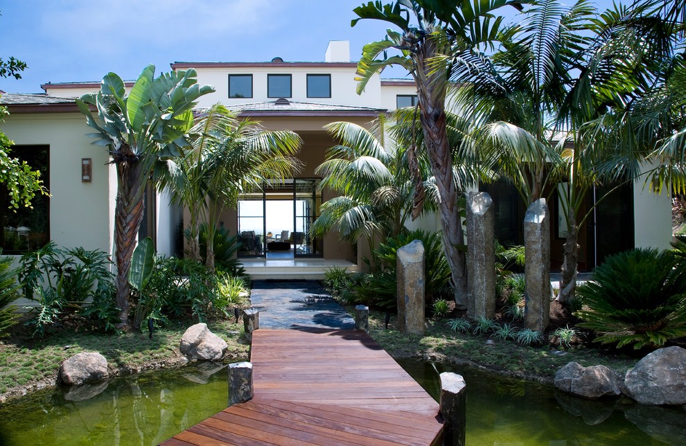 Foto på en stor tropisk trädgård i full sol framför huset, med en damm och trädäck