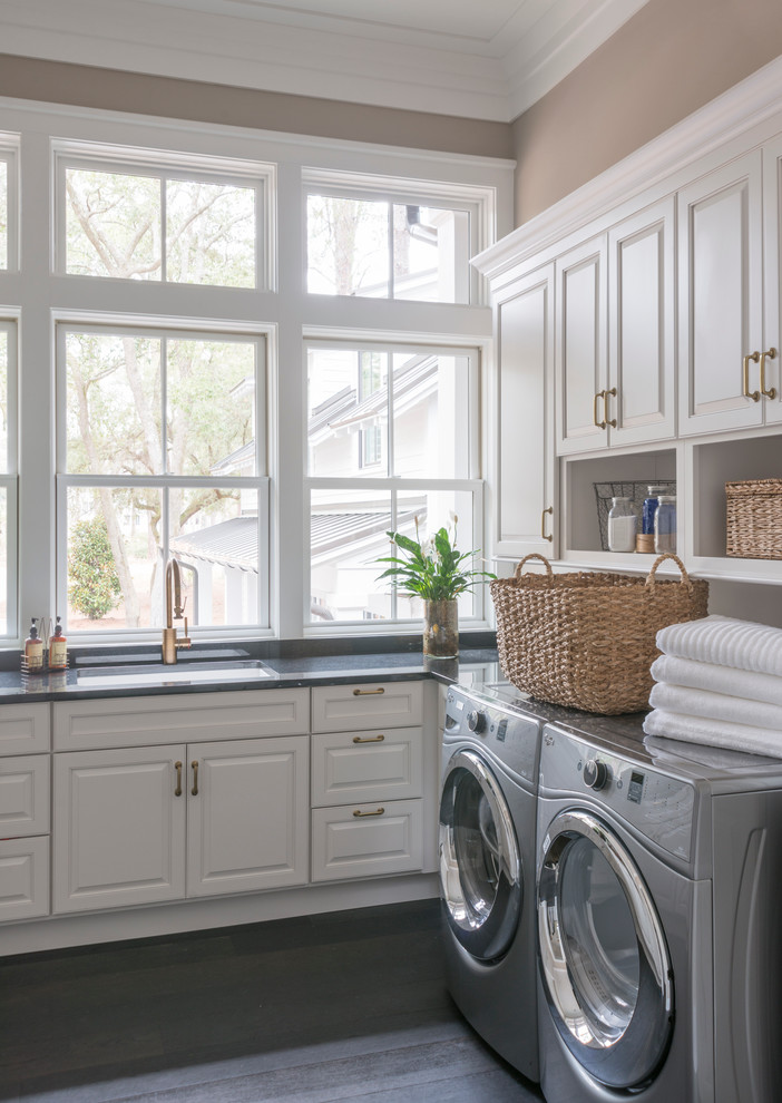This is an example of a large classic l-shaped utility room in Charleston with a submerged sink, raised-panel cabinets, white cabinets, brown walls, dark hardwood flooring, a side by side washer and dryer and black floors.