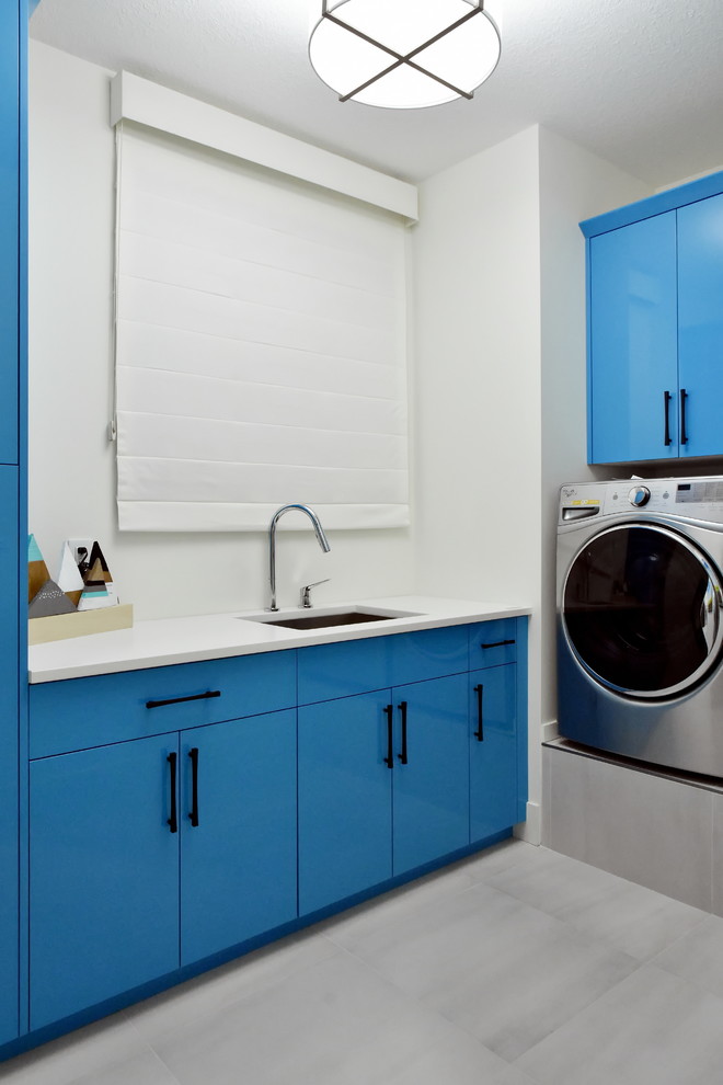 Bild på ett mellanstort funkis l-format grovkök, med en undermonterad diskho, släta luckor, blå skåp, bänkskiva i kvarts, vita väggar och en tvättmaskin och torktumlare bredvid varandra