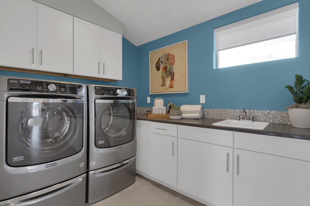 Foto på en stor funkis grå tvättstuga enbart för tvätt, med en nedsänkt diskho, släta luckor, vita skåp, bänkskiva i kvarts, blå väggar, klinkergolv i keramik, en tvättmaskin och torktumlare bredvid varandra och grått golv