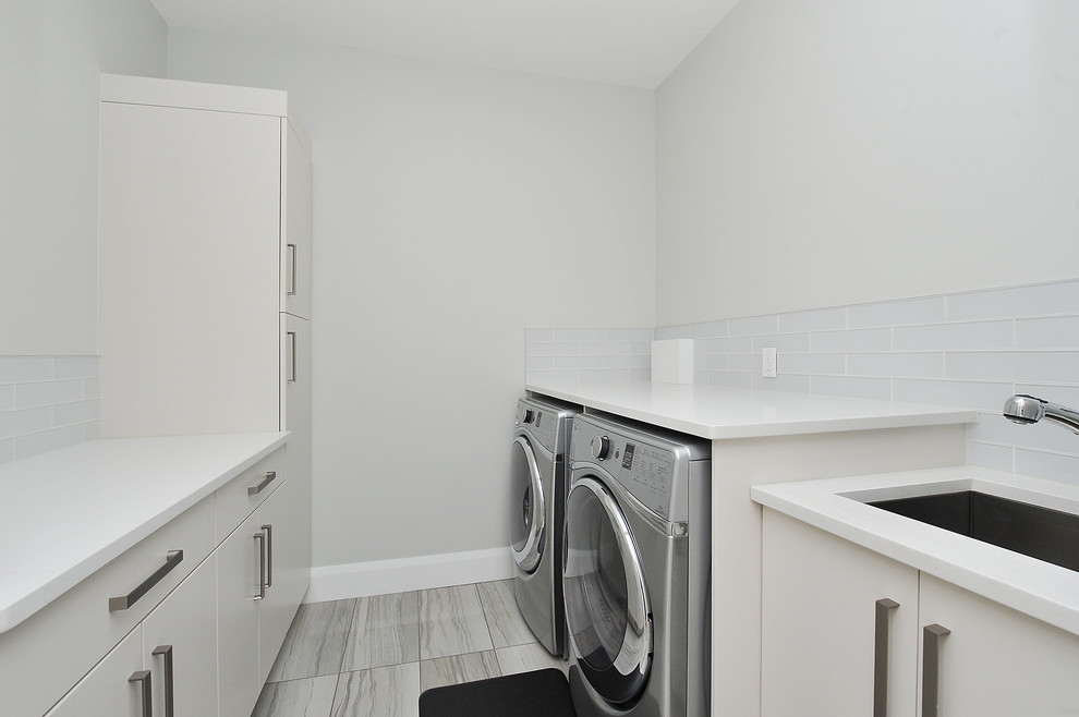 Modern inredning av en mellanstor vita parallell vitt tvättstuga enbart för tvätt, med en undermonterad diskho, släta luckor, vita skåp, bänkskiva i kvartsit, grå väggar, klinkergolv i porslin, en tvättmaskin och torktumlare bredvid varandra och beiget golv