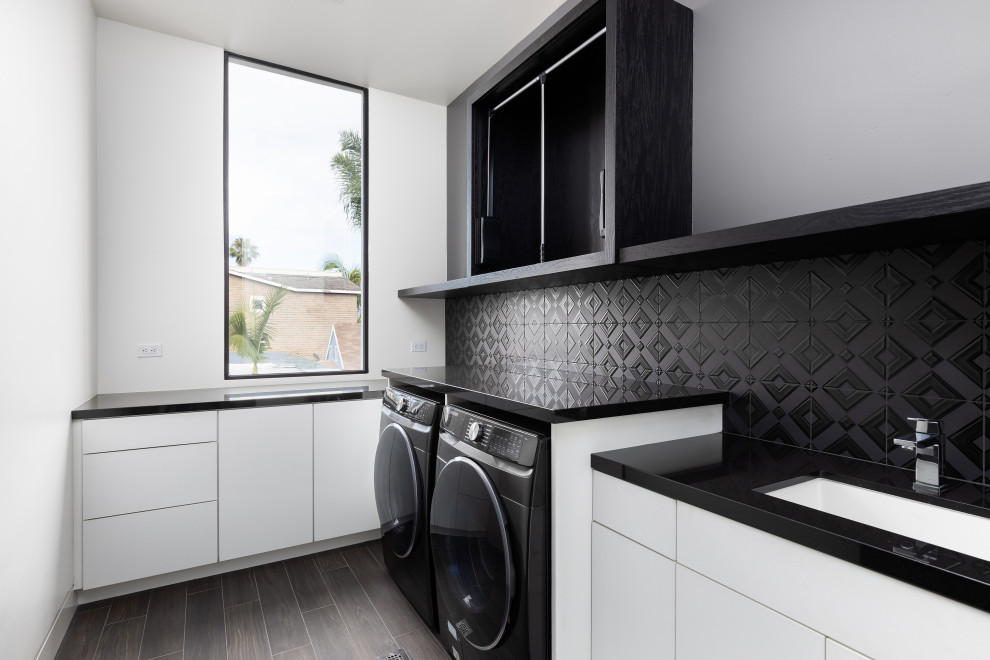 サンディエゴにある小さなコンテンポラリースタイルのおしゃれな洗濯室 (一体型シンク、白いキッチンパネル、白い壁、左右配置の洗濯機・乾燥機、ベージュの床、黒いキッチンカウンター) の写真