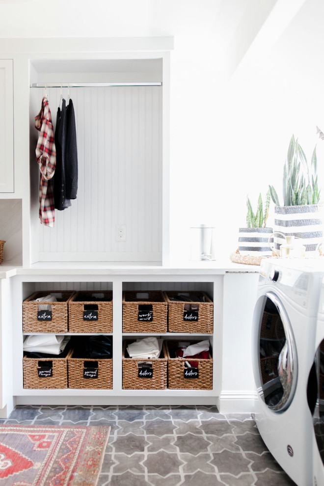 Modelo de cuarto de lavado tradicional renovado con armarios abiertos, puertas de armario blancas, paredes blancas, lavadora y secadora juntas, suelo gris y encimeras blancas
