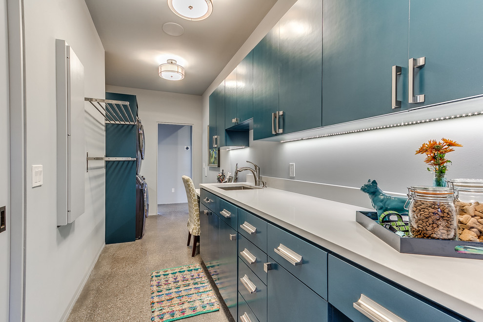 オクラホマシティにある高級な中くらいなコンテンポラリースタイルのおしゃれな家事室 (I型、アンダーカウンターシンク、フラットパネル扉のキャビネット、青いキャビネット、白い壁、コンクリートの床、上下配置の洗濯機・乾燥機、クオーツストーンカウンター、ベージュの床、白いキッチンカウンター) の写真