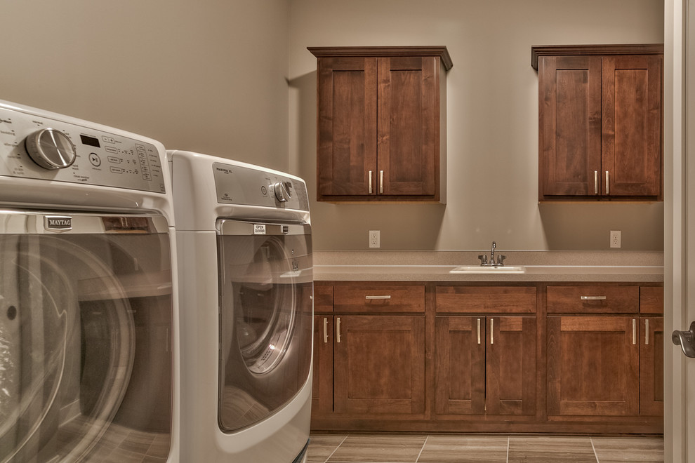 Example of a large tuscan ceramic tile dedicated laundry room design in Omaha with recessed-panel cabinets, medium tone wood cabinets, solid surface countertops, gray walls, a side-by-side washer/dryer and a drop-in sink