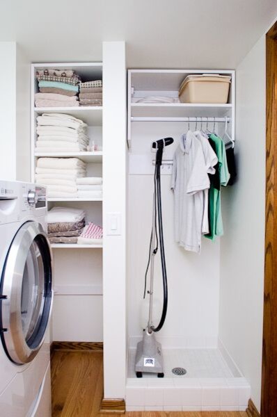Foto på en liten funkis l-formad tvättstuga enbart för tvätt, med vita väggar, ljust trägolv, öppna hyllor, vita skåp och en tvättmaskin och torktumlare bredvid varandra