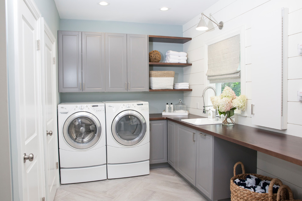 Inredning av en klassisk stor l-formad tvättstuga enbart för tvätt, med luckor med upphöjd panel, grå skåp, träbänkskiva, blå väggar, klinkergolv i porslin, en tvättmaskin och torktumlare bredvid varandra, grått golv och en nedsänkt diskho