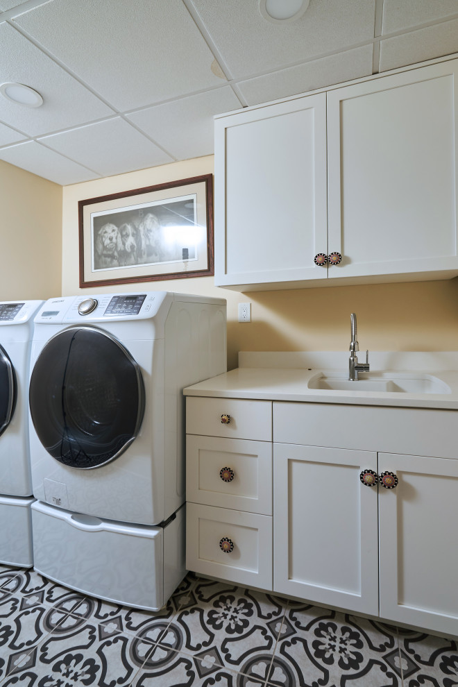 Inredning av en lantlig mellanstor linjär tvättstuga enbart för tvätt, med en integrerad diskho, skåp i shakerstil, vita skåp, bänkskiva i kvarts, beige väggar, betonggolv, en tvättmaskin och torktumlare bredvid varandra och grått golv