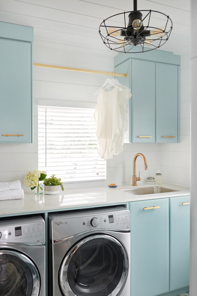 Modelo de cuarto de lavado lineal vintage con fregadero bajoencimera, armarios con paneles lisos, puertas de armario azules, paredes blancas y lavadora y secadora juntas