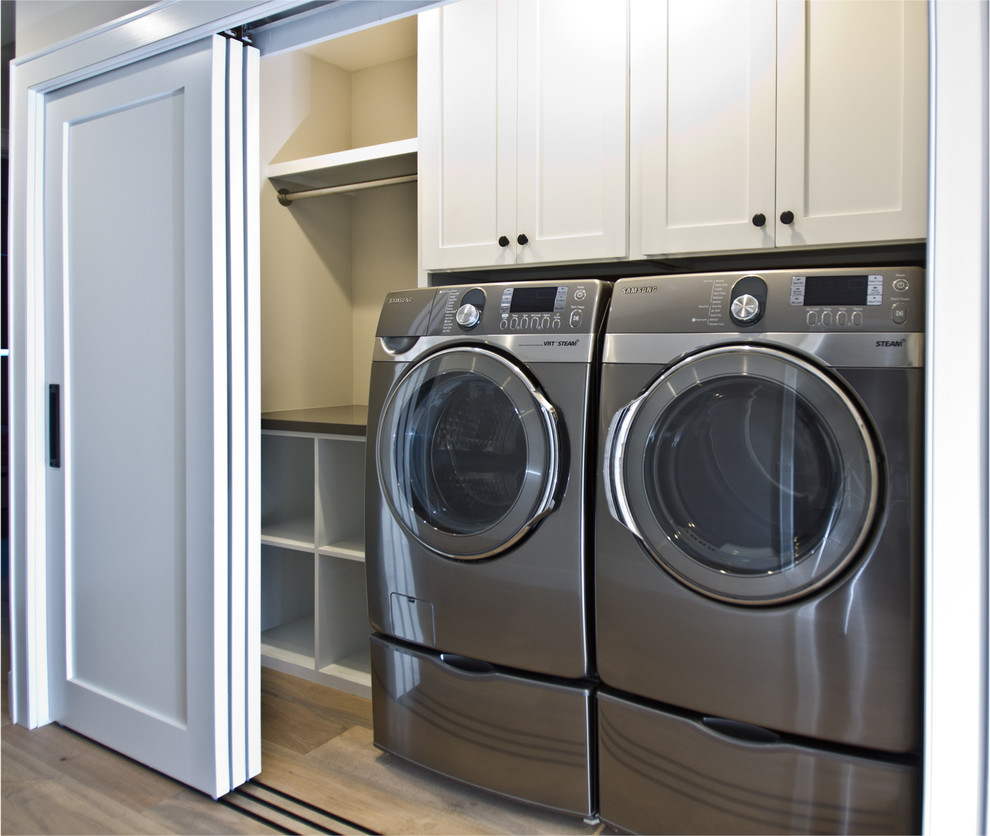 Diseño de armario lavadero lineal clásico pequeño con armarios estilo shaker, puertas de armario blancas, paredes blancas, suelo de madera clara y lavadora y secadora juntas