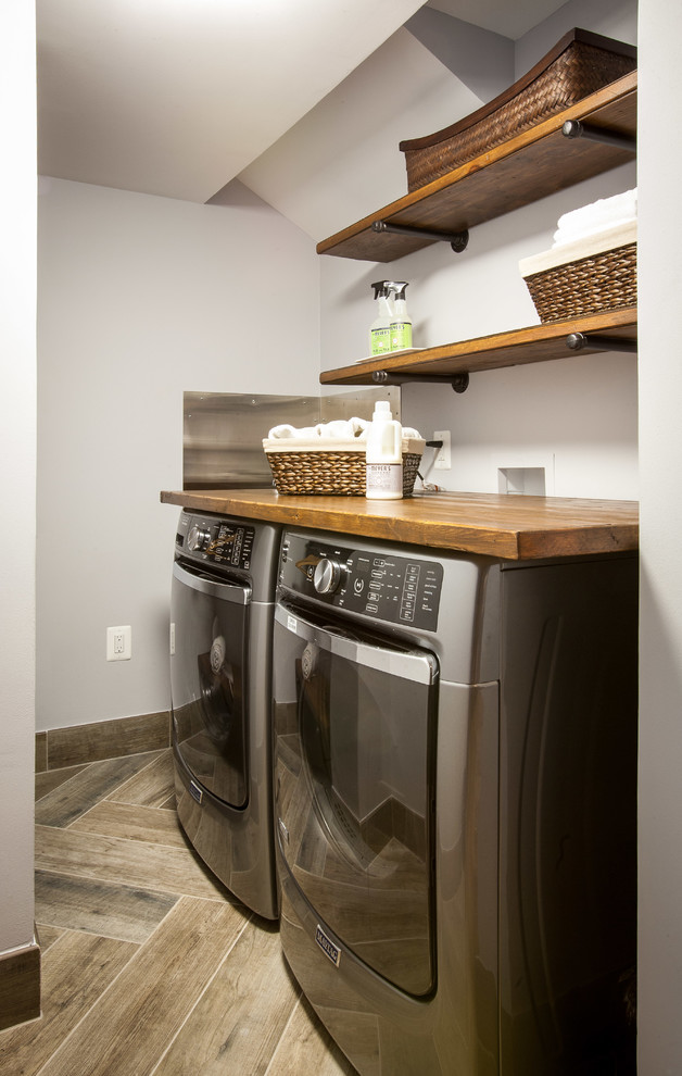 Bild på ett litet rustikt l-format grovkök, med träbänkskiva, grå väggar, klinkergolv i porslin, en tvättmaskin och torktumlare bredvid varandra och brunt golv