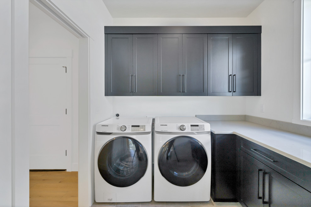 Große Moderne Waschküche in L-Form mit Schrankfronten im Shaker-Stil, schwarzen Schränken, Quarzwerkstein-Arbeitsplatte, weißer Wandfarbe, Waschmaschine und Trockner nebeneinander, grauem Boden und schwarzer Arbeitsplatte in Sonstige