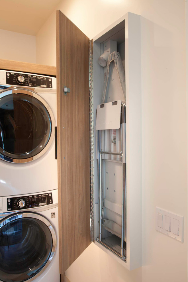 Photo of a small nautical l-shaped separated utility room in San Diego with a submerged sink, flat-panel cabinets, light wood cabinets, engineered stone countertops, blue splashback, cement tile splashback, white walls, porcelain flooring, a stacked washer and dryer, beige floors and white worktops.