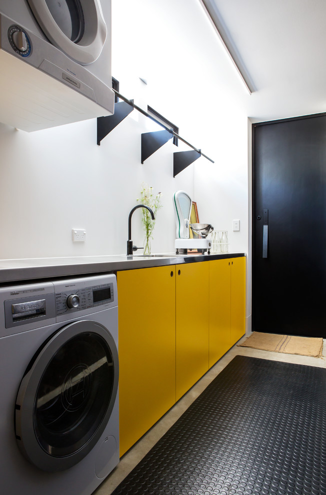 Immagine di una sala lavanderia contemporanea con lavello sottopiano, ante lisce, ante gialle, pareti bianche, pavimento in cemento, lavatrice e asciugatrice a colonna, pavimento grigio e top grigio