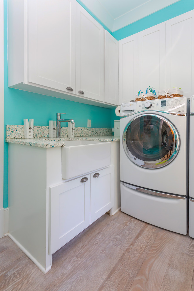 This is an example of a medium sized beach style galley utility room in Charleston with a belfast sink, raised-panel cabinets, white cabinets, recycled glass countertops, blue walls, light hardwood flooring, a side by side washer and dryer, beige floors and multicoloured worktops.