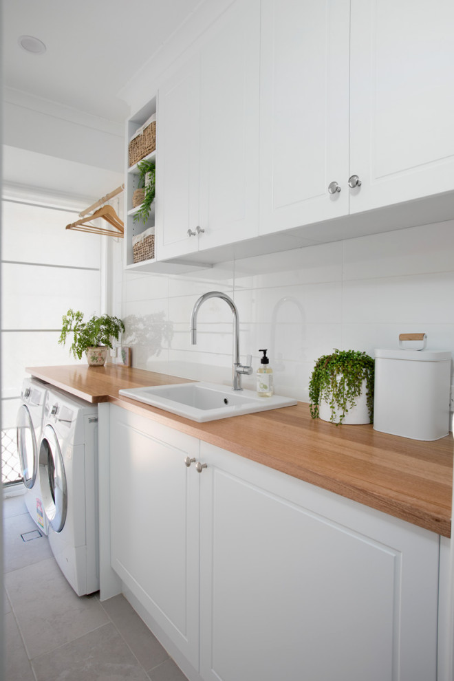 Waschküche mit Waschbecken, Schrankfronten im Shaker-Stil, weißen Schränken, Arbeitsplatte aus Holz, weißer Wandfarbe, Waschmaschine und Trockner nebeneinander und grauem Boden in Brisbane
