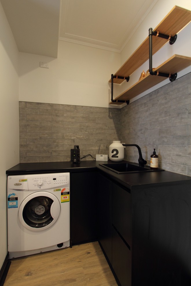 Foto på en liten industriell l-formad tvättstuga enbart för tvätt, med en enkel diskho, svarta skåp, laminatbänkskiva, grå väggar, laminatgolv, en tvättpelare och släta luckor