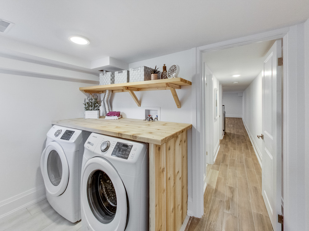Minimalistisk inredning av en mellanstor linjär tvättstuga enbart för tvätt, med öppna hyllor, skåp i ljust trä, träbänkskiva, vita väggar, en tvättmaskin och torktumlare bredvid varandra, klinkergolv i keramik och grått golv