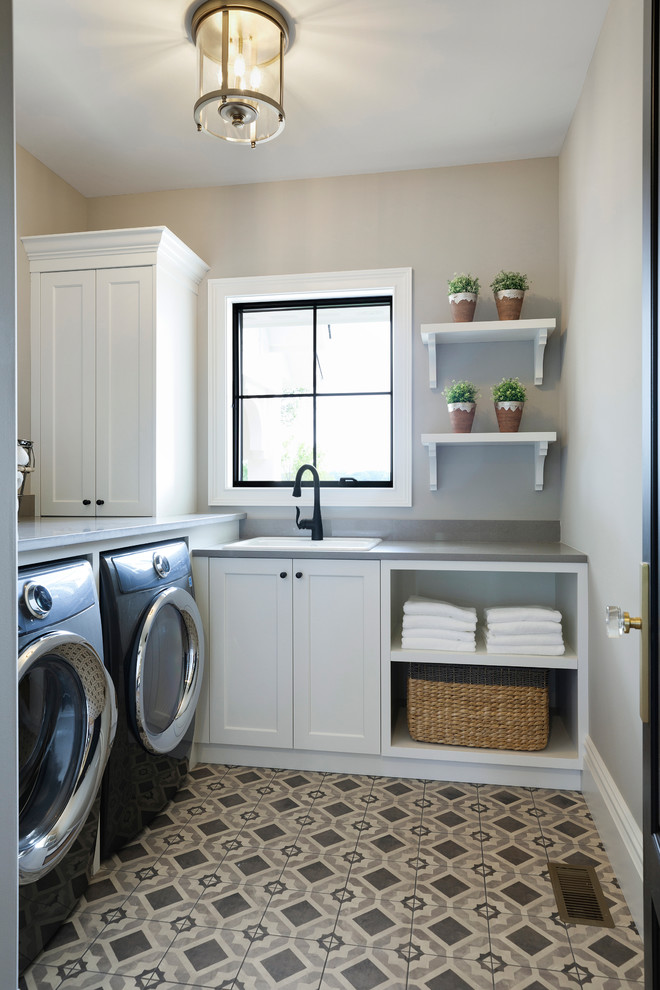 Inredning av en stor l-formad tvättstuga enbart för tvätt, med vita skåp, grå väggar, klinkergolv i porslin, en tvättmaskin och torktumlare bredvid varandra och flerfärgat golv