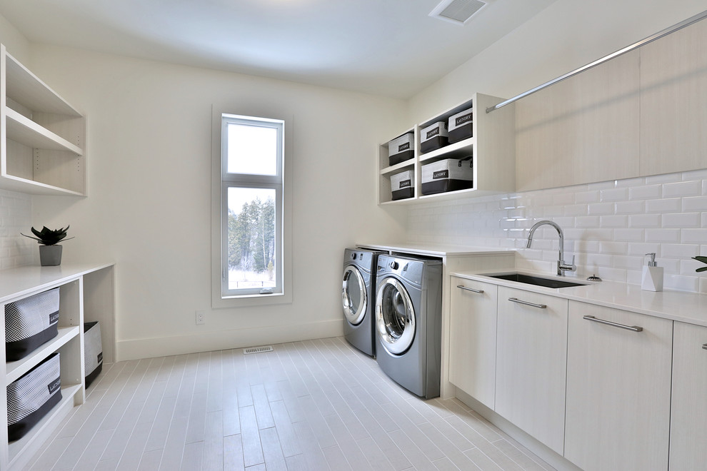 Multifunktionaler, Zweizeiliger, Großer Moderner Hauswirtschaftsraum mit Waschbecken, flächenbündigen Schrankfronten, hellen Holzschränken, Quarzwerkstein-Arbeitsplatte, weißer Wandfarbe, hellem Holzboden und Waschmaschine und Trockner nebeneinander in Toronto