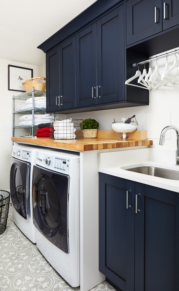 Immagine di una sala lavanderia tradizionale con lavello sottopiano, ante in stile shaker, ante blu, top in legno, pareti bianche, lavatrice e asciugatrice affiancate, pavimento grigio e top bianco