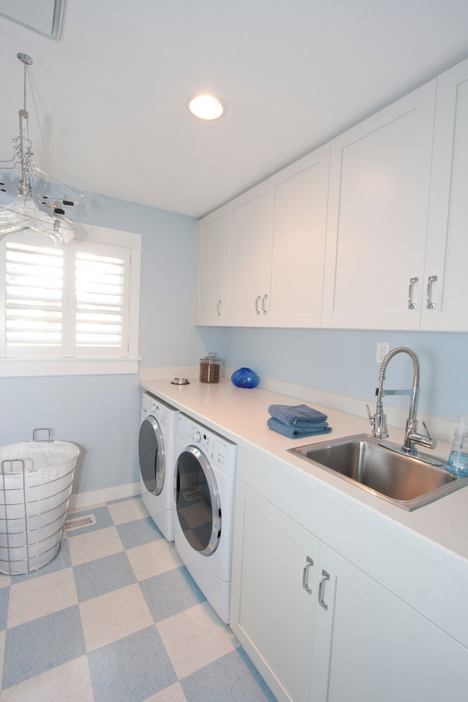 Foto på en stor vintage vita linjär tvättstuga enbart för tvätt, med laminatbänkskiva, en nedsänkt diskho, vita skåp, skåp i shakerstil, blå väggar, linoleumgolv, en tvättmaskin och torktumlare bredvid varandra och flerfärgat golv
